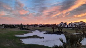 A golf course with sand bunkers and palm trees, surrounded by buildings with a sunset in the background.