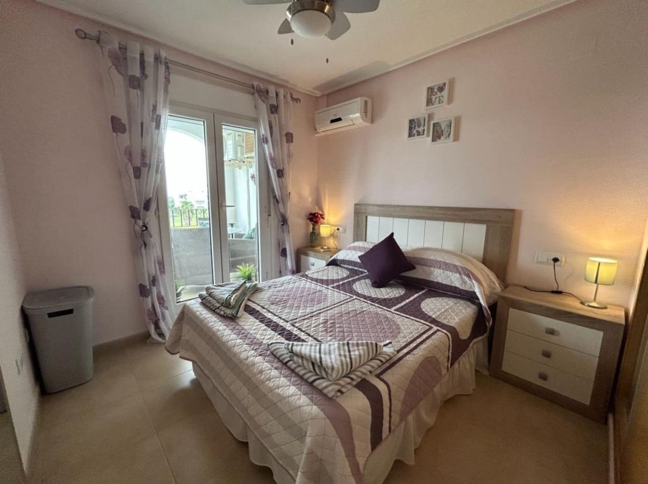 Une chambre à coucher avec un lit, une table de chevet, une lampe, une poubelle et une fenêtre avec des rideaux.