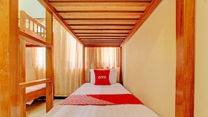 Ein schmales Hotelzimmer mit Etagenbett, einem Einzelbett mit rotem Kissen und einer rot-weißen Decke mit Muster.