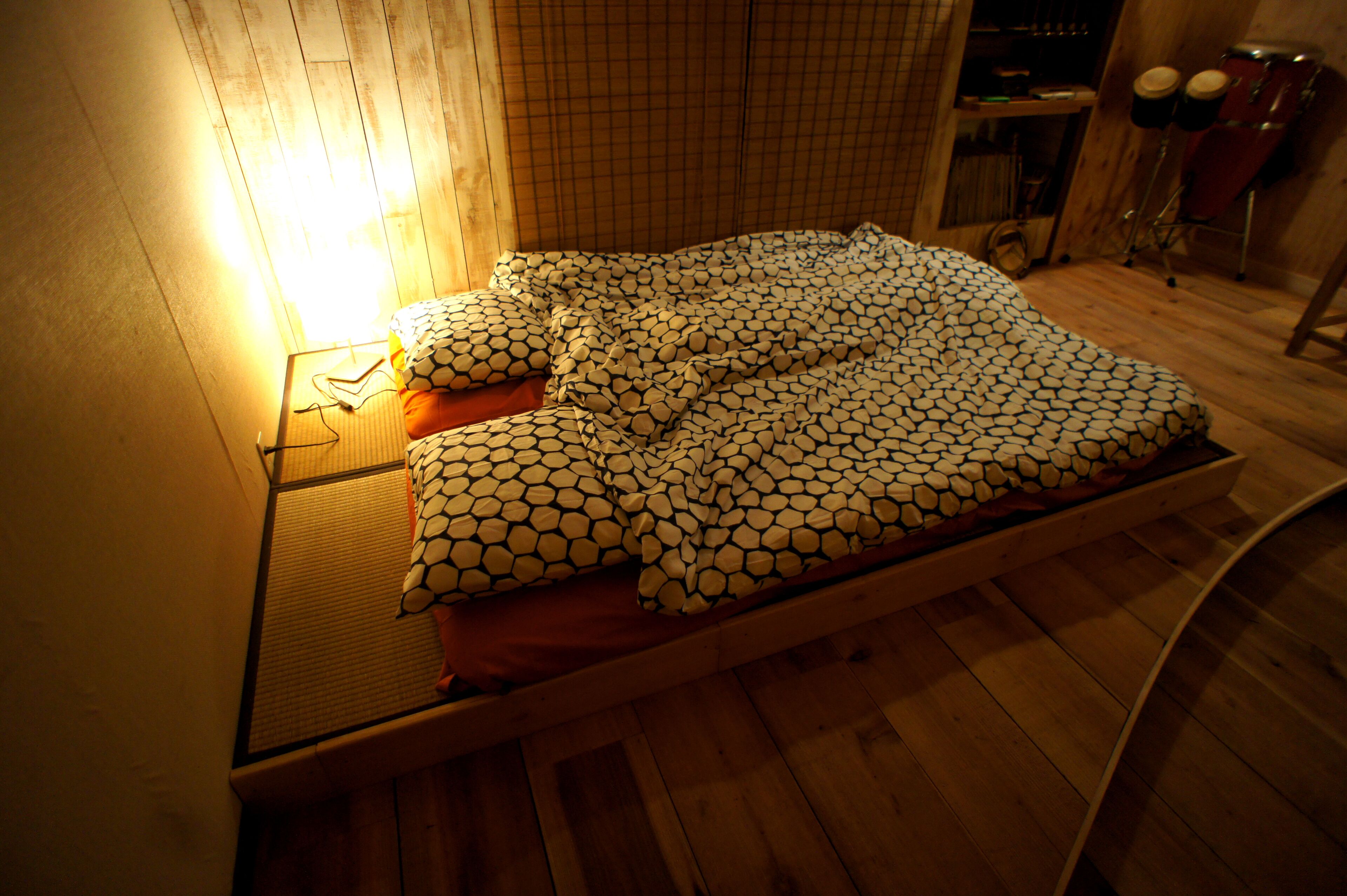 A bed with a patterned blanket, a bedside table with a lamp, and a wooden floor.