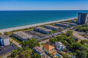 An aerial view of a coastal hotel with multiple buildings, a parking lot, and a beach in the background.
