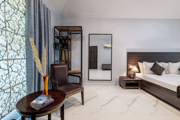 A hotel room with a bed, a chair, a small table with a book, a mirror, and a nightstand with a lamp. -  Valley By Grassfield (Viratnagar)