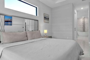 A modern bedroom with a large bed, a nightstand, a lamp, a painting, and a bathroom with a bathtub.