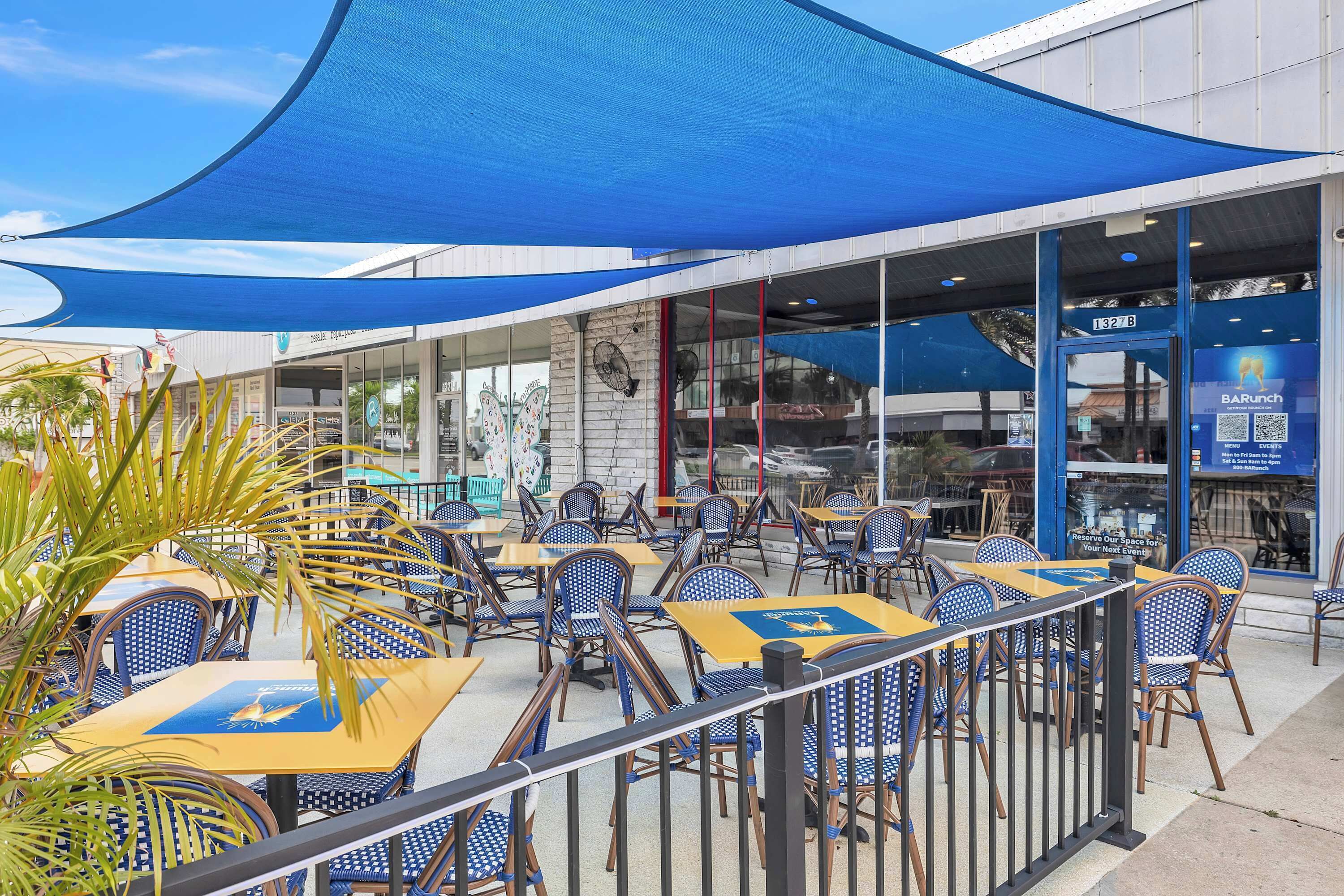 An outdoor seating area with tables and chairs, a blue awning, and a storefront with signage.