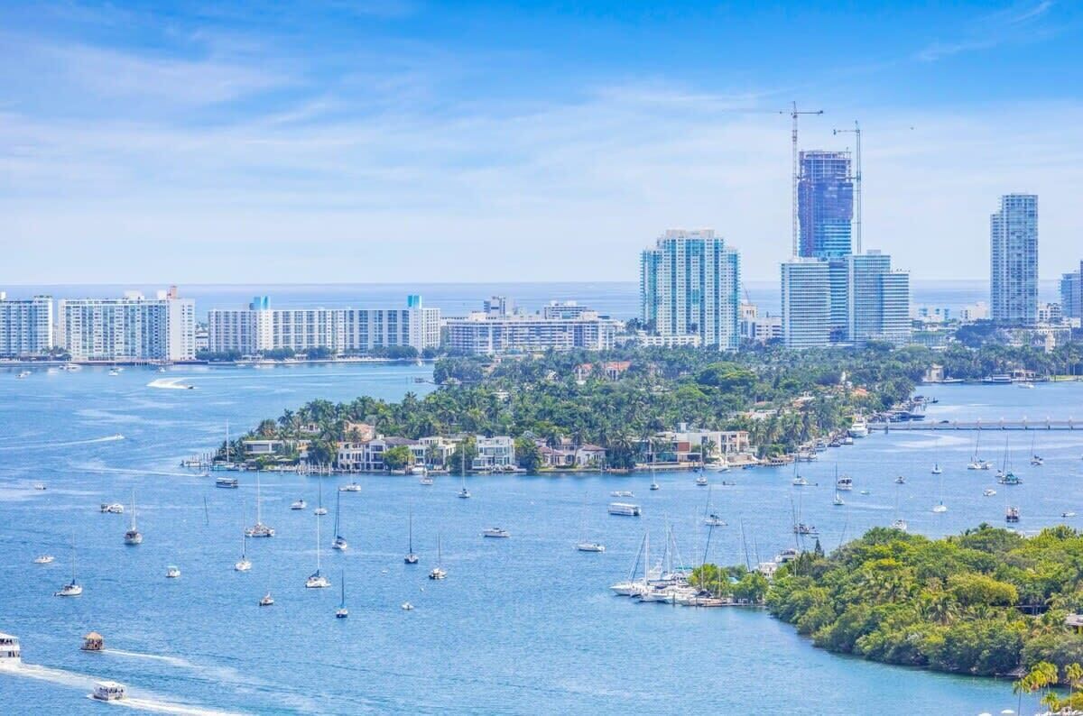 Paisagem costeira com uma marina repleta de barcos, edifícios altos e uma ilha densamente vegetada.