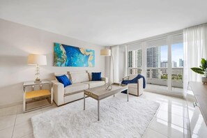 A modern living room with a white sofa, a coffee table, and a large window offering a city view. - Bay Overlooking Balcony & Full Kitchen | StayCozy (Miami)