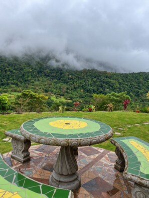 Un tavolo rotondo con decorazioni in mosaico, circondato da un giardino ricco di piante e affaccio su una collina boscosa.