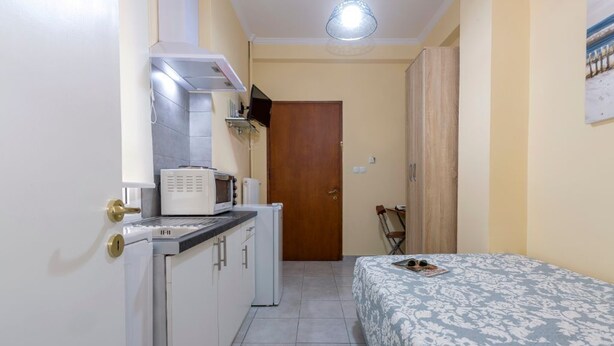 Cocina compacta con microondas, tostadora y refrigerador pequeño. Se ve una cama con colcha estampada en la habitación contigua.