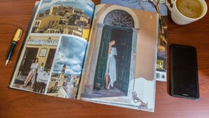 Una revista abierta a una página con una vista urbana, una mujer con un vestido blanco y una taza de café sobre una mesa de madera.