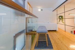 A modern hotel room with a wooden floor, a bed with white linens, a small stool, a wall-mounted plant, and a glass partition.