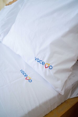 Un oreiller blanc orné du logo « Vacedo » brodé en bleu et en rouge, posé sur un lit.