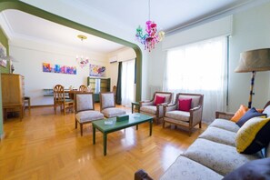 Un salon spacieux doté d’un parquet, d’une table basse verte et d’un canapé agrémenté de coussins colorés. Au fond, on aperçoit un coin repas avec une table et des chaises.