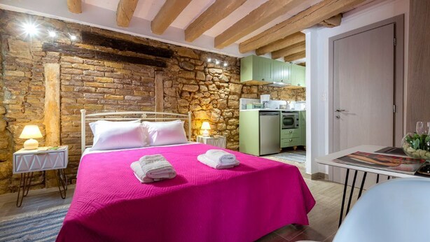 Una stanza con parete in pietra, un letto con copriletto rosa, un piccolo tavolo con una lampada e un angolo cottura sullo sfondo.