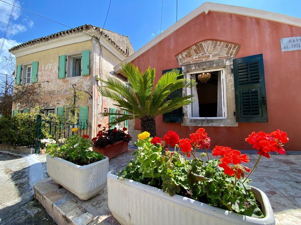 A building with a pink facade, green shutters, and a stone entrance. There are flower beds with red and yellow flowers in front.