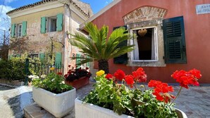 A building with a pink facade, green shutters, and a stone entrance. There are flower beds with red and yellow flowers in front. - The Old Oven House - The apartment was renovated to strict traditional standards. (Corfu)
