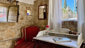 A room with stone walls, a red chair, a marble table with a book, and a window with a view of greenery. - The Old Oven House - The apartment was renovated to strict traditional standards. (Corfu)
