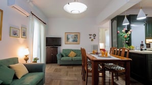 A living room with a green sofa, a dining table with chairs, a TV, and artwork on the walls. - Pausa Verde - Recently renovated apt, private parking (Corfu)