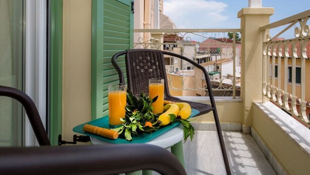 A balcony with a table set for breakfast, including a chair, a tray with bananas, a bottle of orange juice, and a glass of orange juice. - Pausa Verde - Recently renovated apt, private parking (Corfu)