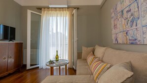 A living room with a sofa, a television on a wooden cabinet, a small round table with a bottle and glasses, and a large window with sheer curtains. - Le Boulevard - stylish, renovated apartment (Corfu)