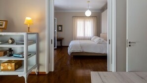A bedroom with a wooden floor, a bed with white bedding, a nightstand with a lamp, and a bookshelf with books and a clock. - Le Boulevard - stylish, renovated apartment (Corfu)