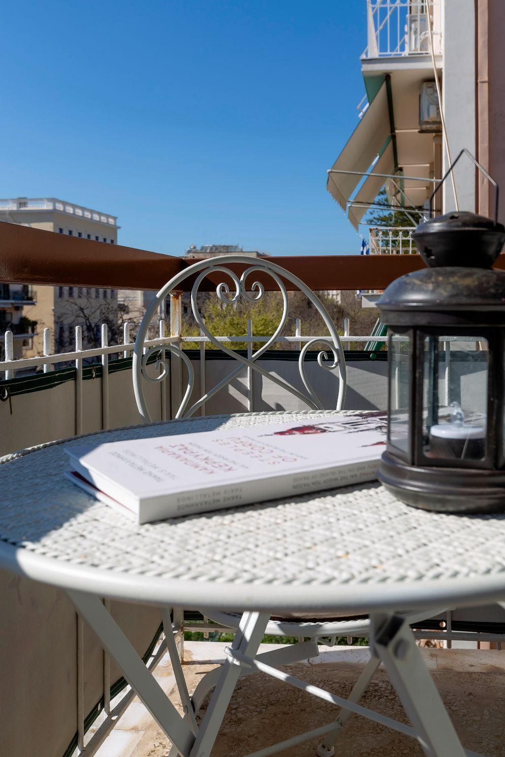 A white metal table with a book and a lantern on it, set on a balcony with a clear view of buildings in the background.