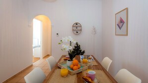 Et spiseområde med et træbord, hvide stole, en vase med hvide blomster, appelsiner og en tallerken med en croissant.