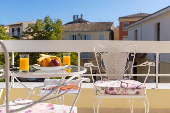 Un balcón con una mesa dispuesta para el desayuno, que incluye frutas y jugo, y una silla con un cojín estampado de flores.