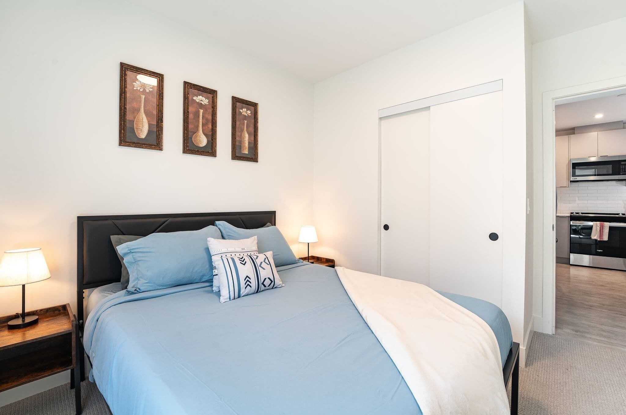 A bedroom with a bed, bedside lamps, a door, and a view into the kitchen.