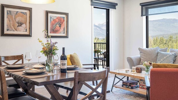 A dining area with a table set for four, a sofa, and a view of mountains through a window. - Revytalize - Cozy 3 Bedroom Condo (Revelstoke)