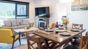 A modern living room with a sofa, coffee table, dining table, chairs, a fireplace, a flat-screen TV, and a painting on the wall. - Revytalize - Cozy 3 Bedroom Condo (Revelstoke)