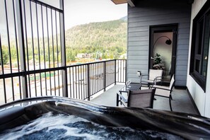 A balcony with a hot tub, outdoor seating, and a view of a forested area.
