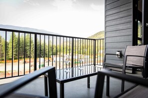Un balcon avec une table et des chaises, donnant sur une zone boisée avec des montagnes à l’horizon.