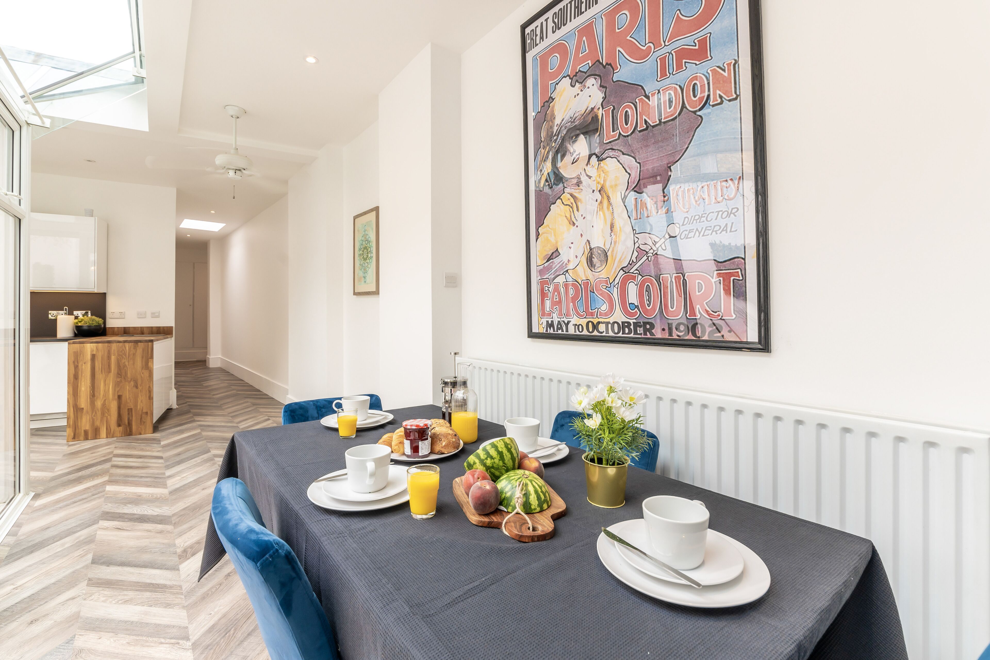 A dining area with a table set for breakfast, including fruits and beverages, and a poster on the wall.