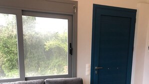 Um sofá cinzento com uma mesa de centro preta, uma porta azul e uma janela com vista para árvores.