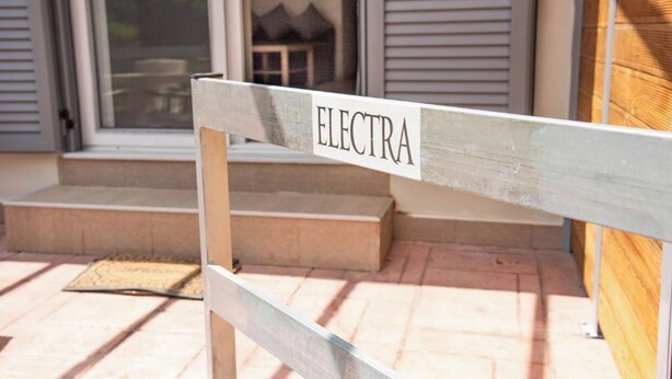 Un banc en bois sur lequel est inscrit le mot « ELECTRA », placé devant une maison qui a une porte et des marches menant à un porche.