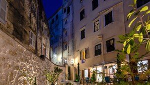 A narrow street with buildings on either side, some with shutters, and a few outdoor seating areas with tables and chairs. - Volto Achilles Suite (Corfu)
