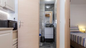 Ein kleines Zimmer mit einer Küchenzeile an einer Seite, einem Badezimmer mit Spiegel und Waschbecken sowie einem Bett mit gestreifter Bettwäsche auf der anderen Seite.