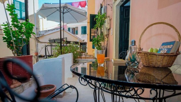 A glass table with a wicker basket and a glass of orange juice on a balcony with potted plants and a view of buildings. - Campiello Blue Studio - Central cozy studio with garden in Corfu old town (Corfu)