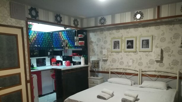 A bedroom with a bed, a nightstand, and a bedside table. There is a kitchen area with a refrigerator, a sink, and a stove. The room has patterned wallpaper and a door with a decorative frame. - Pento Studio - Ground floor apt, located near Corfu square (Kerkira)