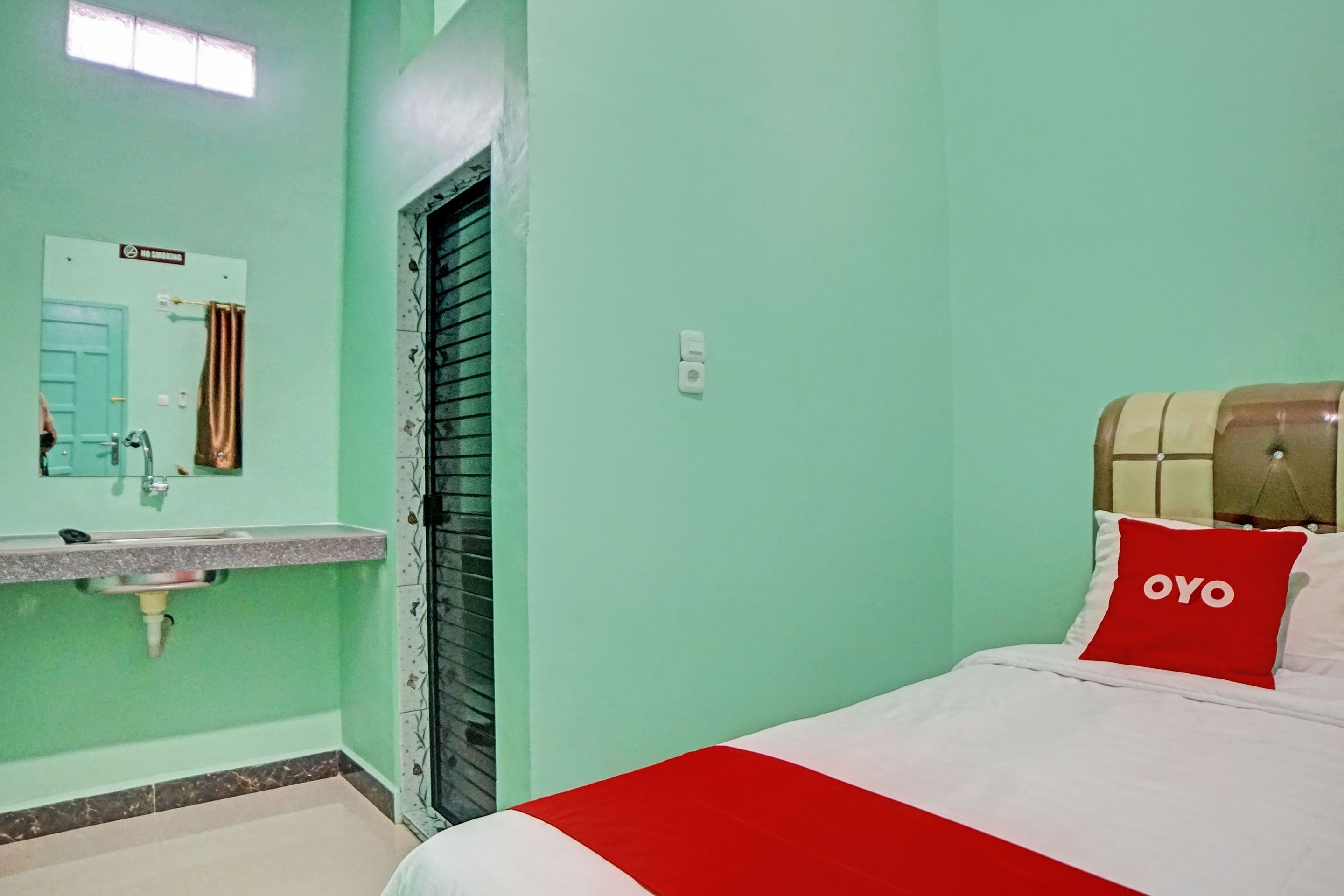 Une chambre avec un lit, une couvre-lit rouge et blanche, un oreiller rouge orné du mot « OYO », une salle de bain avec un lavabo et un miroir, et une porte avec une pancarte.