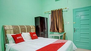 Una cama bien hecha con una colcha roja y blanca, dos almohadas rojas con el texto 'OYO', un armario de color oscuro y una puerta verde.