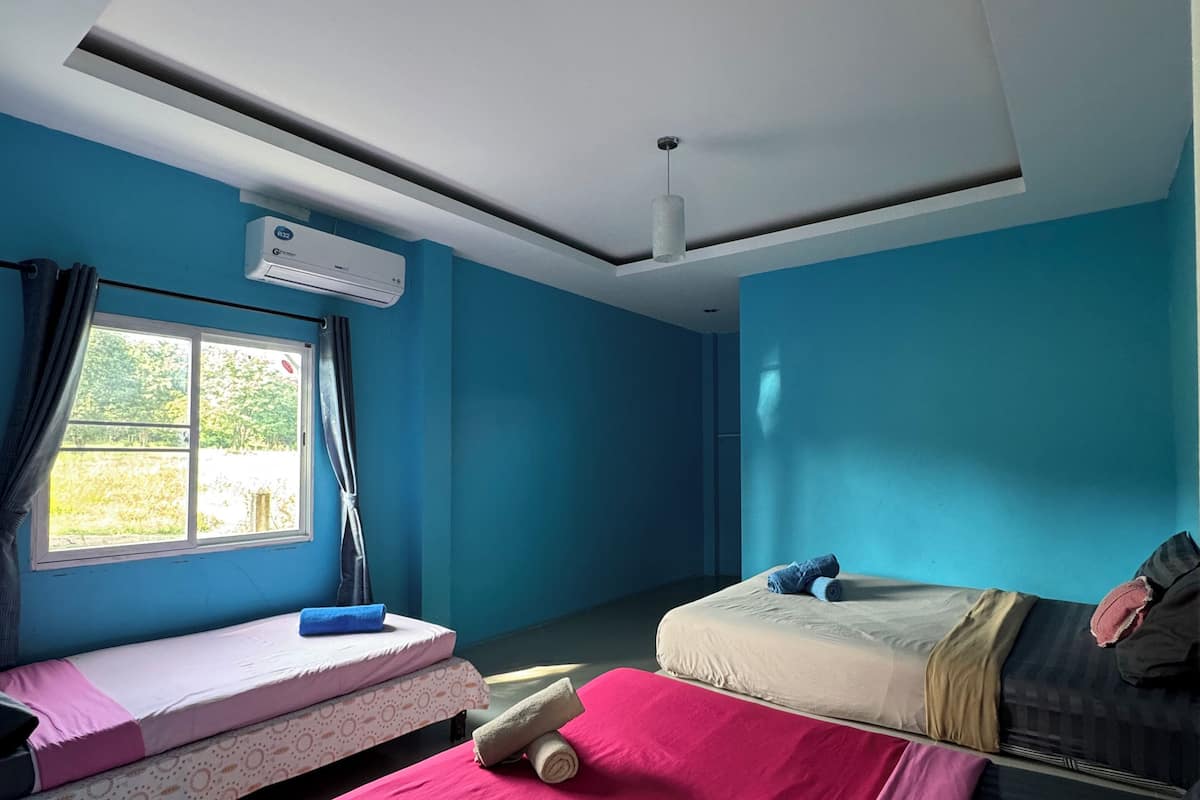 Quarto com duas camas, uma com colcha vermelha e outra com colcha bege. Há uma janela com cortinas, um ar condicionado e um ventilador de teto.
