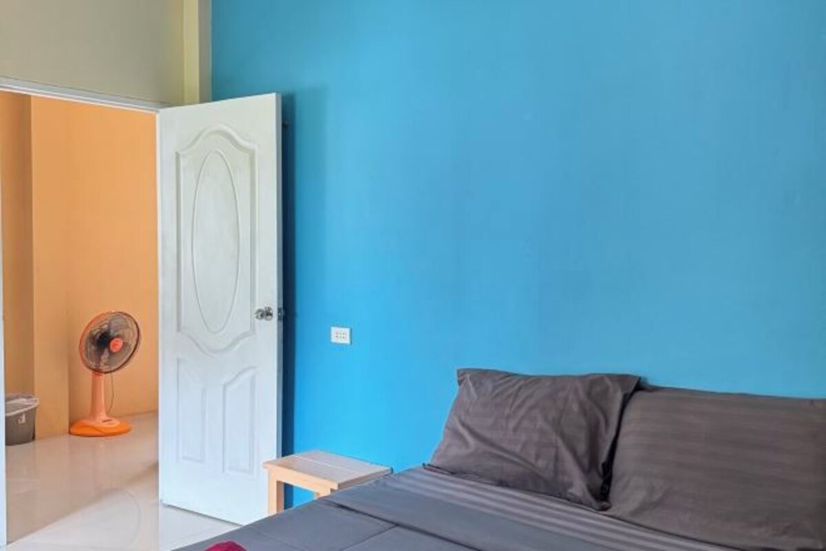 A room with a bed, a small table, a fan, and a door.