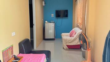 A hotel room with a bed, a desk with a chair, a TV mounted on the wall, a refrigerator, and a fan.
