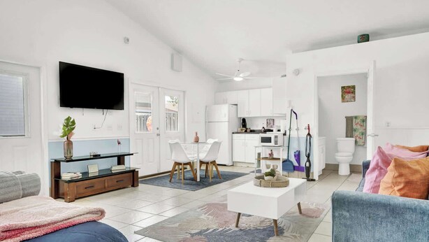 Una sala de estar moderna con televisor de pantalla plana, un comedor con sillas blancas, una cocina con armarios blancos y un baño con inodoro y ducha.