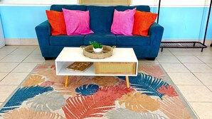 Un sofá azul con cojines rosa y naranja, una mesita blanca con una cesta trenzada y un libro, y una alfombra colorida con estampado de hojas.