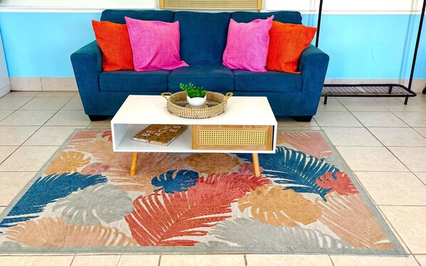Un canapé bleu agrémenté de coussins roses et orange, une table d’appoint blanche avec un panier en osier et un livre, et un tapis aux motifs colorés représentant des feuilles.