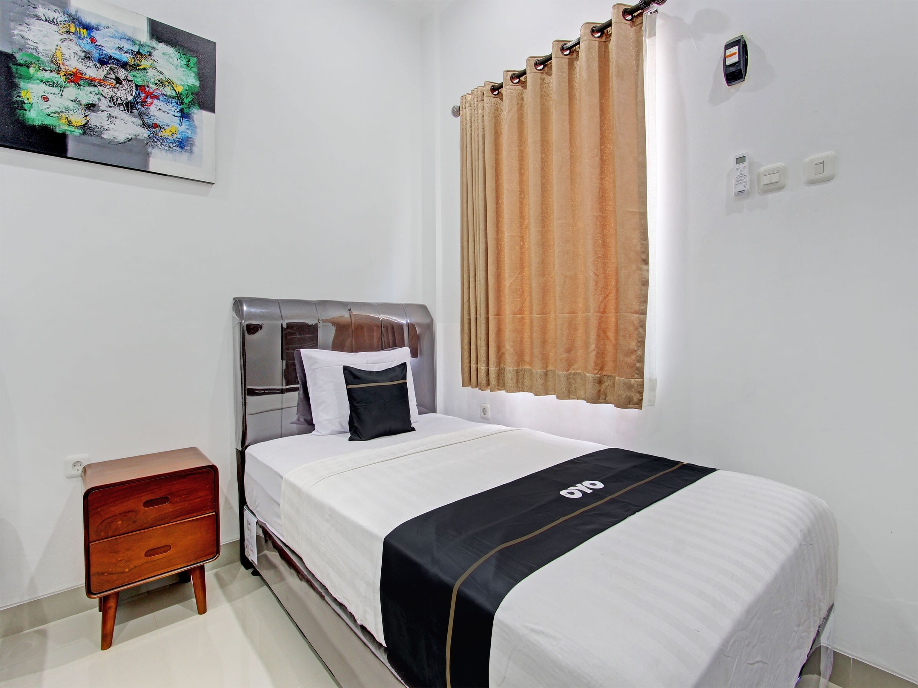 A neatly arranged bedroom with a bed, a bedside table, a wall-mounted light switch, and a window with curtains.