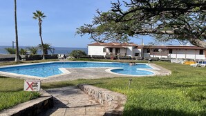 A swimming pool with a stone deck, surrounded by grass and palm trees, with a view of the sea and buildings in the background. - Tamara Alto (Acantilados de Los Gigantes)