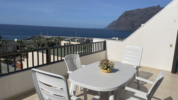 A balcony with a table and chairs, overlooking a coastal area with buildings and a mountain in the distance. - Tamara Alto (Acantilados de Los Gigantes)